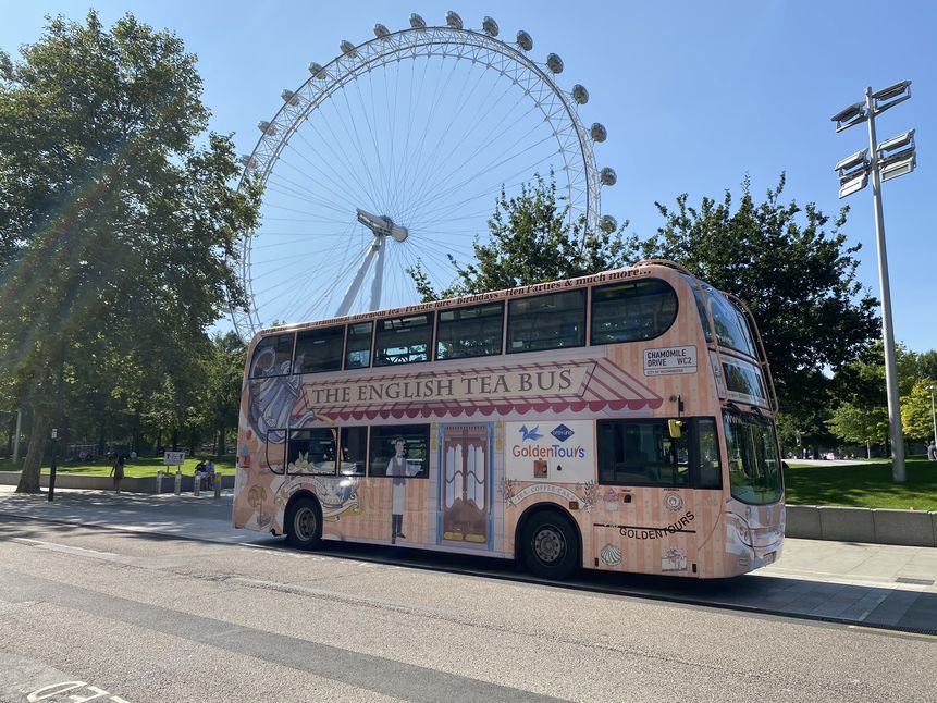  A peach-colored, double-decker bus is parked on a street in London. The bus is decorated with the words "THE ENGLISH TEA BUS" and "Goldentours," along with illustrations of teacups, cakes, and other afternoon tea items. In the background, the London Eye is clearly visible on a sunny day. The bus appears to be part of a tour that combines afternoon tea with a panoramic tour of the city.