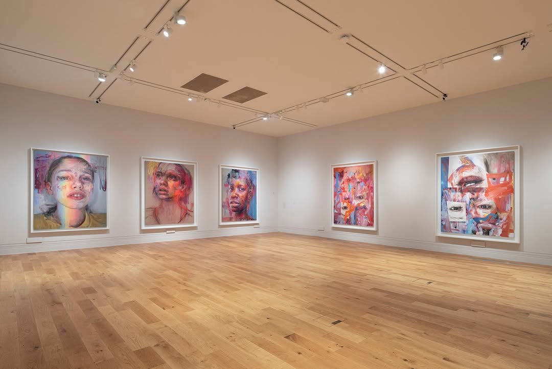 The image shows an art gallery with a light wooden floor and white walls. Five large, colorful paintings, possibly by artist Jenny Saville, are hung on one wall. Each painting features a portrait, with distorted and abstract elements. The first painting on the left is a close-up of a person's face, the second and third show more stylized portraits, and the last two are highly abstract with bold brushstrokes and multiple facial features overlapping. 